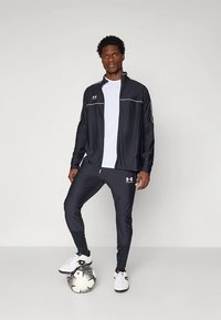 Svart atletisk treningsjakke og bukser med hvite detaljer, kombinert med en hvit t-skjorte og svarte treningssko, stående på en fotball.