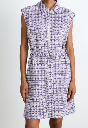 Robe sans manches à carreaux violets et blancs avec col, fermeture éclair à l'avant et ceinture assortie en tissu avec boucle circulaire, portée par une femme debout.