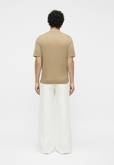 Άτομο με σγουρά μαλλιά που φοράει μπεζ κοντομάνικο πουκάμισο και άσπρο παντελόνι φαρδιάς γραμμής, στέκεται με την πλάτη προς ένα απλό φόντο.