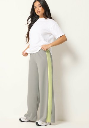 Kvinde iført løse grå bukser med gule sidelængder, en simpel hvid oversize t-shirt og hvide sneakers, stående op ad en lys væg.