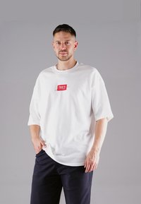 White oversized t-shirt in cotton with a round neck and short sleeves, featuring a red rectangular logo on the front. Dark pants.