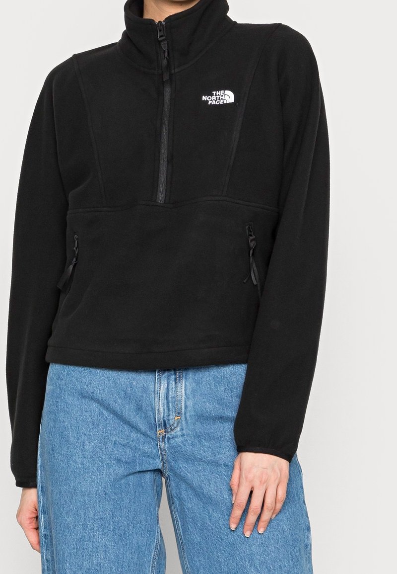 Musta fleecetunika korkealla kauluksella, jossa neljännesvetoketju ja kaksi vetoketjullista taskua, pehmeä tekstuuri ja rintamuksessa logo.