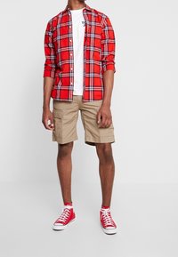 Camicia a quadri rossa con accenti neri e bianchi, t-shirt bianca, pantaloni cargo beige fin sulla ginocchia e sneaker rosse alte.