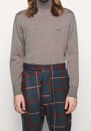 Person trägt einen beigen Rollkragenpullover mit kleinem Logo und eine hoch Taillierte karierte Hose mit Knöpfen am Bund.