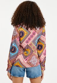 Mønstret, farverig skjorte med knapper, der har forskellige geometriske designs, med lange ærmer og en buet kant, parret med denimshorts.