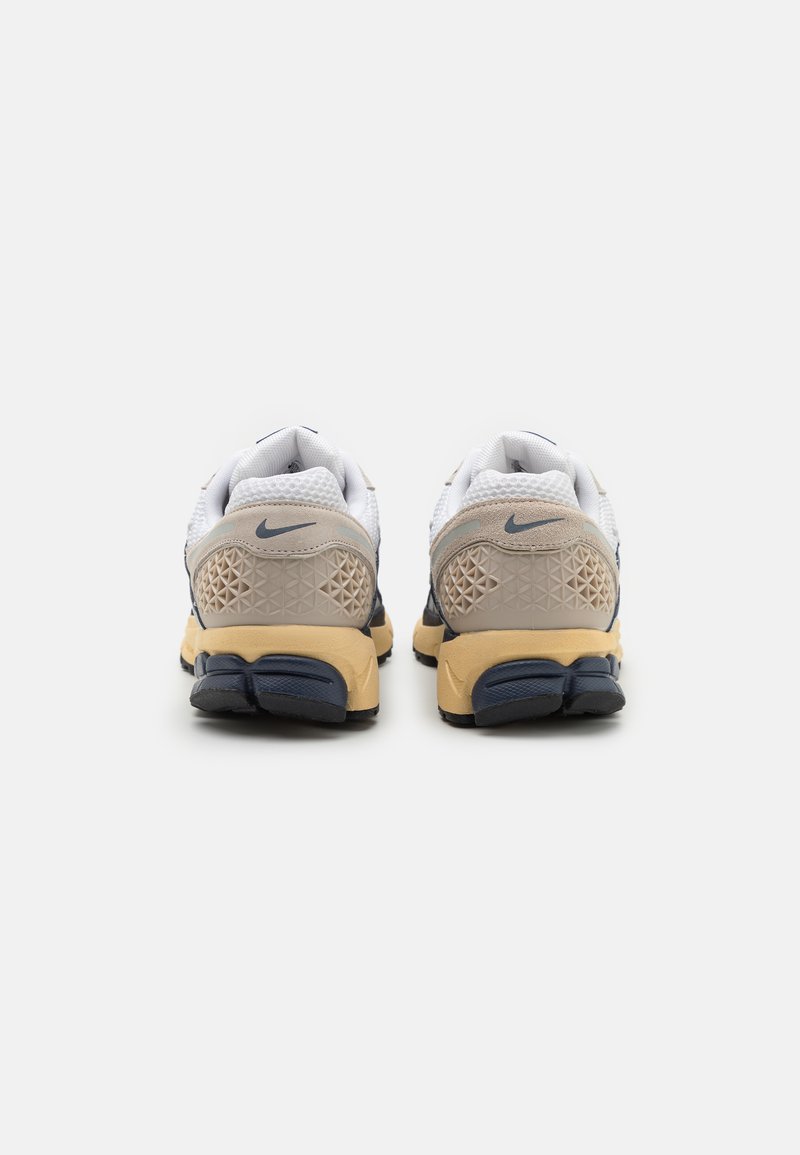 Zapatillas Nike con una parte superior de malla blanca, acentos en beige y azul marino, talón texturizado y una suela de goma con una unidad de aire visible.