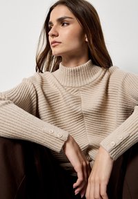 Jersey de punto de canalé beige con un cuello alto, que presenta un diseño texturizado y detalles de botones en los puños. Tejido suave y acogedor.