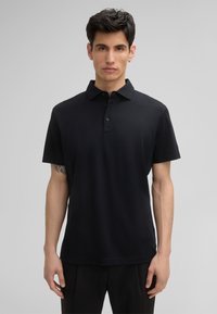 Czarny polo wykonany z gładkiego materiału, z kołnierzykiem i zapięciem na trzy guziki. Krótkie rękawy i luźny fason.
