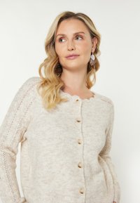 Cardigan lavorato a maglia color beige chiaro con collo scallop, dotato di cinque bottoni rotondi e dettagli in maglia aperta sulle maniche.