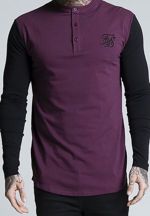 Mężczyzna w koszulce henley z długim rękawem, purpurowym korpusem, czarnymi rękawami, trzema guzikami oraz małym, czarnym, wyszywanym logo na piersi.
