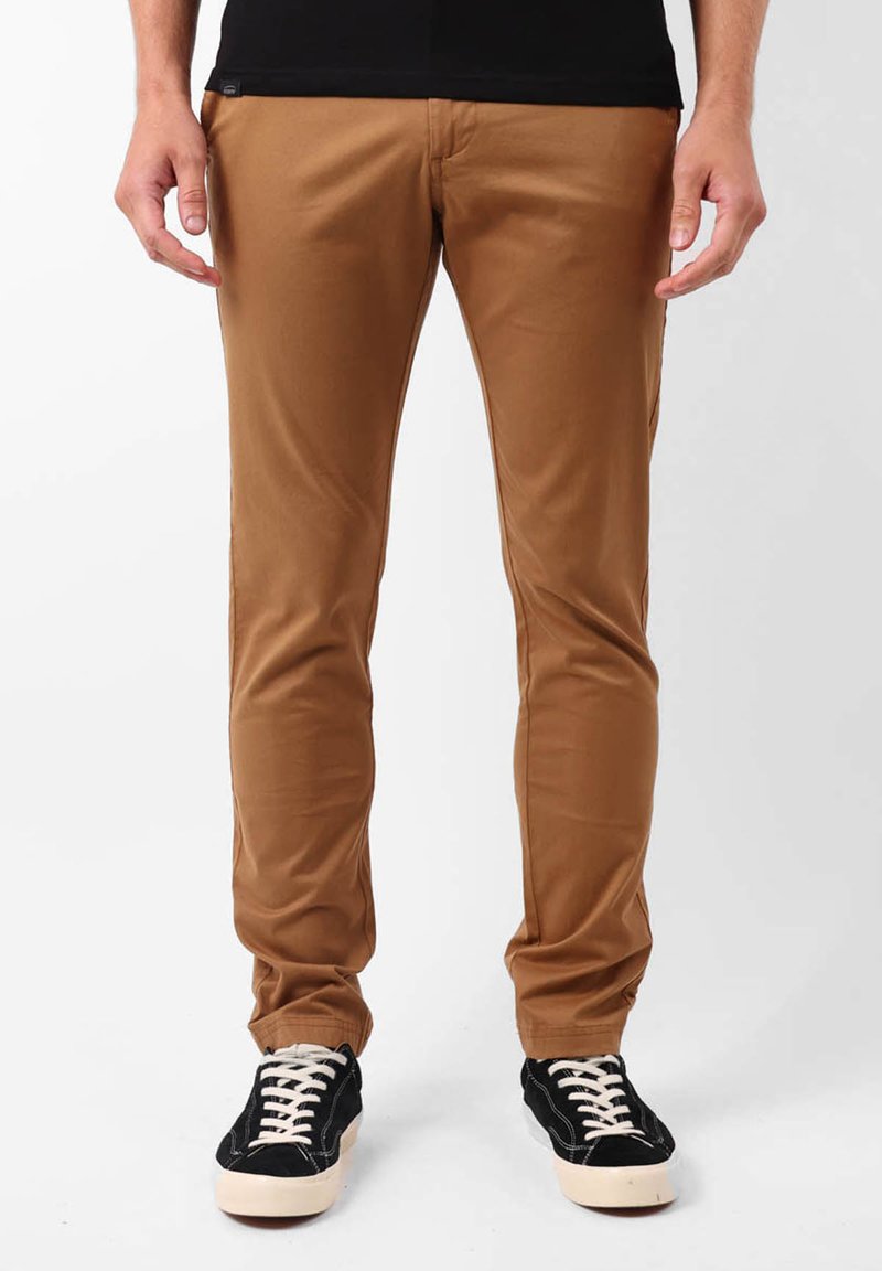 Pantalons en mélange de coton beige avec une coupe slim, présentant une texture lisse et un design classique à cinq poches. Associés à des baskets noires.