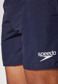 Shorts de bain bleus marine en matériau léger avec un logo Speedo blanc sur la jambe inférieure et une texture lisse. Pas de motifs supplémentaires.