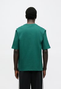 Zelené bavlnené tričko s okrúhlym výstrihom a krátkymi rukávmi, s voľným strihom a jemnou textúrou, pohľad zo zadnej strany. Jednoduchý dizajn bez ozdôb.