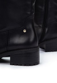 Bota negra de cuero hasta el tobillo con un acabado texturizado, puntera redonda, un tacón bajo y apilado, y un pequeño detalle dorado cerca de la costura. Cierre con cremallera lateral.