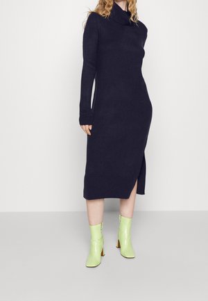 Robe tricotée marine avec un col montant, des manches longues et une fente latérale. Assortie à des bottes chevilles vertes brillantes et un fond minimaliste.