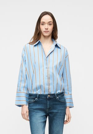 Kobieta ubrana w jasnoniebieską koszulę na guziki z brązowo-białymi pionowymi paskami, w połączeniu z niebieskimi dżinsami, stojąca na jednolitym tle.