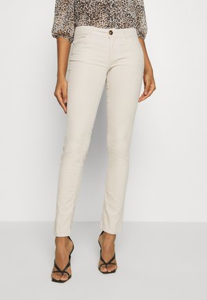 Kvinna iförd beige slim-fit byxor, leopardmönstrad blus och svarta remsandaler som står mot en enkel vit bakgrund.