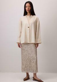 Jachetă bej din in cu un singur nasture auriu, asortată cu o fustă argintie cu paiete și pantofi maro cu talpă joasă. Design simplu și curat.