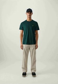 Dark green cotton t-shirt with short sleeves and a small yellow logo, paired with beige cargo pants and black sneakers.