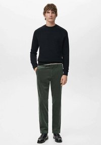 Mężczyzna w czarnym swetrze z okrągłym dekoltem, ciemnozielonych spodniach sztruksowych i czarnych eleganckich butach, stojący z jedną ręką w kieszeni na jednolitym tle.