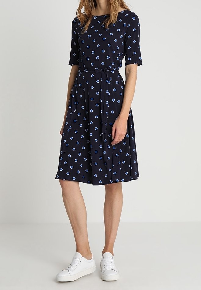 Marinblå klänning med korta ärmar och A-linjeformad silhuett, med ett blått cirkelmönster och en knuten midja, kombinerad med vita sneakers.