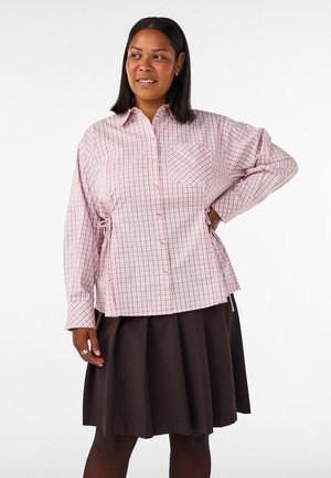 Donna con carnagione media che indossa una camicia rosa a quadri con bottoni e una gonna scura plissettata, in piedi con una mano sui fianchi.