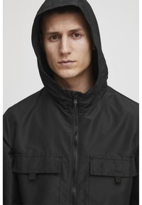 Črna zip-up jakna s kapuco, ki ima dva sprednja žepa in teksturirano tkanino. Oblikovanje je minimalistično in utilitarno.