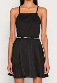 Svart klänning med spagettiband, åtsittande bodice och utsvängd kjol. Har Calvin Klein Jeans-logotyp vid midjan. Slät tygetextur.