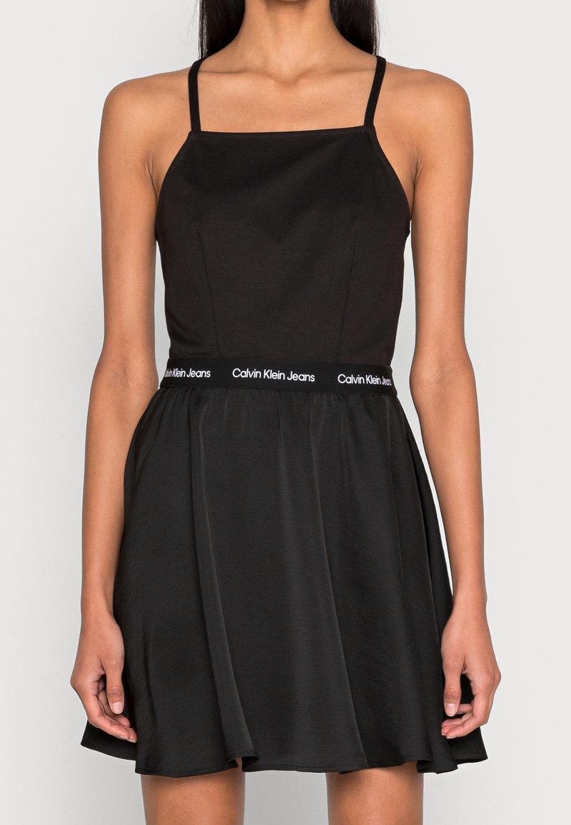 Svart klänning med spagettiband, åtsittande bodice och utsvängd kjol. Har Calvin Klein Jeans-logotyp vid midjan. Slät tygetextur.