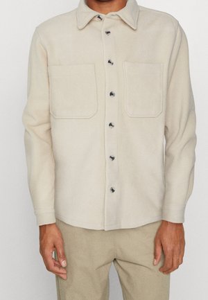 Mand iført en beige fleeceskjorte med knapper og to brystlommer samt beige bukser mod en ensfarvet baggrund.