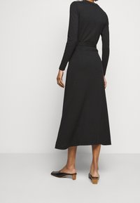 Abito nero a maniche lunghe con cintura in vita, forma ad A, tessuto testurizzato e lunghezza a metà polpaccio. Indossato con mules con tacco aperto.