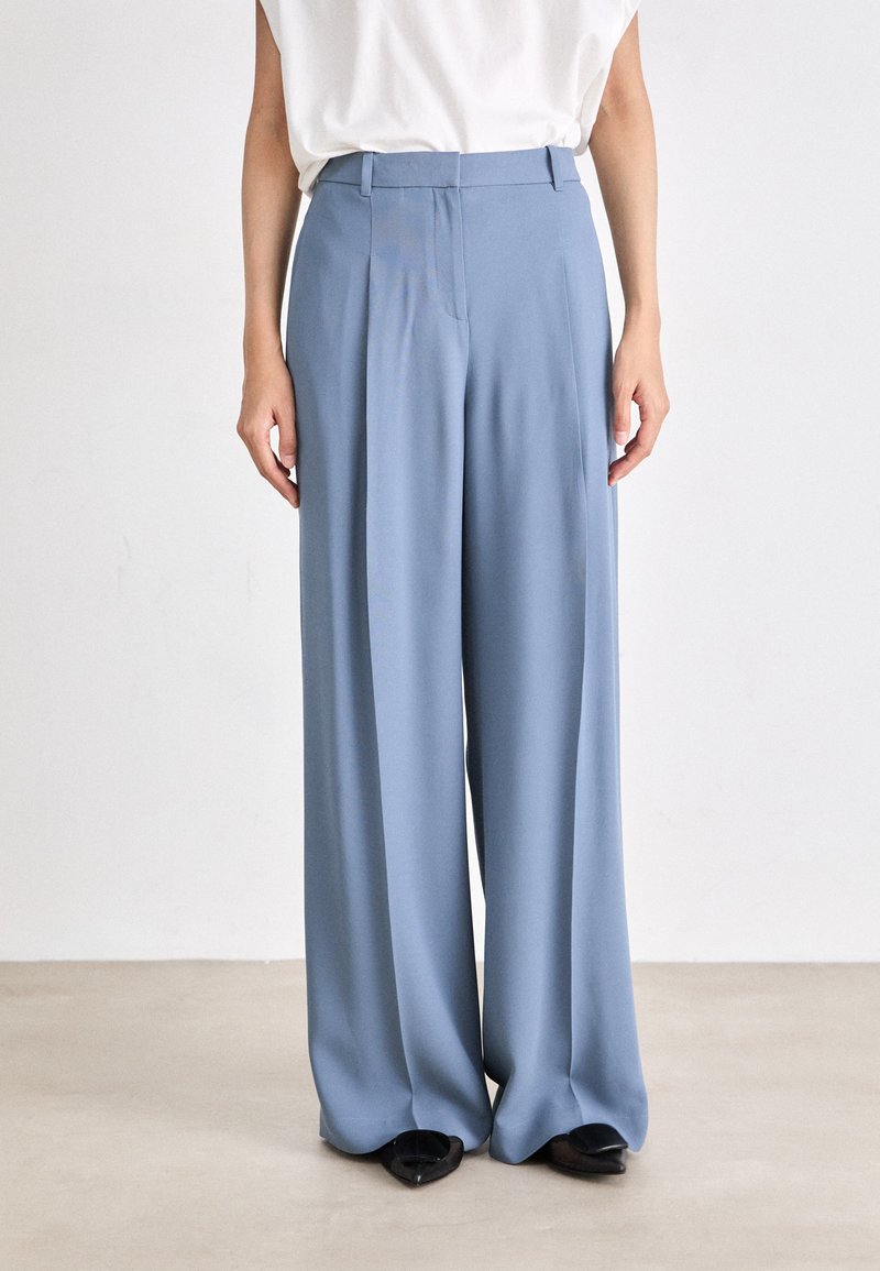 Pantalons larges bleu clair en tissu lisse, présentant une coupe ajustée, des plis devant et des poches latérales discrètes.
