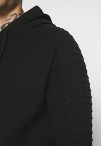 Man som bär en svart ribbad hoodie med texturerade ärmdetaljer, där en del av nacktatuerings syns mot en enfärgad bakgrund.