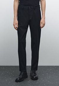 Czarne spodnie z tkaniny, o dopasowanym kroju, z zapięciem na guzik, szlufkami na pasek i bocznymi kieszeniami. W połączeniu z czarnymi sznurowanymi butami na ciemnej podłodze.