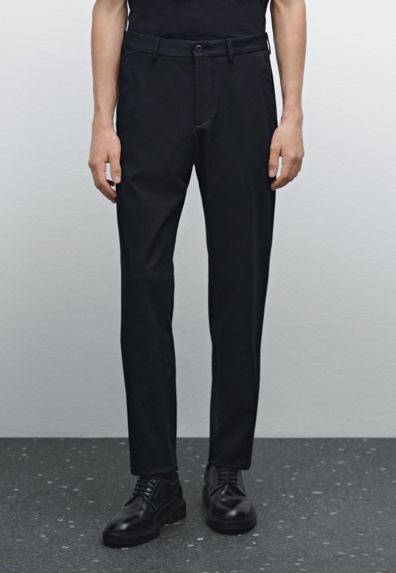 Czarne spodnie z tkaniny, o dopasowanym kroju, z zapięciem na guzik, szlufkami na pasek i bocznymi kieszeniami. W połączeniu z czarnymi sznurowanymi butami na ciemnej podłodze.