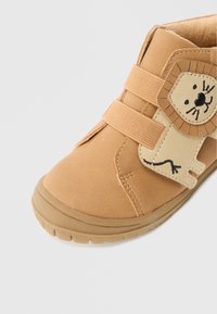 Brūna velcro aizdare apava ar lauvu uzšuvumu un melniem izšuvumiem. Mīksts augšējais materiāls ar gumijas zoli; noapaļota purngala forma.