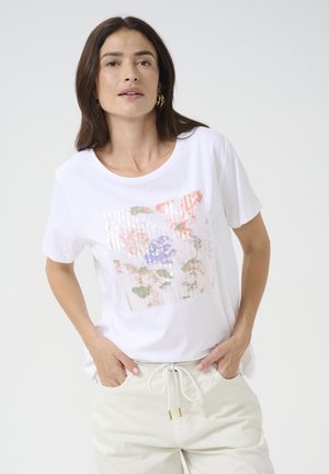 White t-shirt featuring a floral sequin design in pink, green, and blue. Crew neck, short sleeves, relaxed fit, paired with light pants.