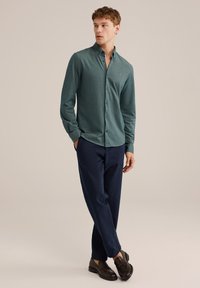 Majica z dolgimi rokavi in gumbi iz zeleno-modre tkanine, kombinirana z mornarsko modrimi hlačami in črnimi loaferji, stoječa na nevtralnem ozadju.