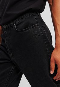 Vaqueros de mezclilla negros con un diseño ajustado, que presentan un sutil logo en relieve en el bolsillo, costuras visibles y una textura suave.
