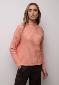 Pull à manches longues côtelé couleur corail clair, doté d'un col montant et d'un tissu texturé. Associé à un pantalon foncé, il offre une coupe décontractée.