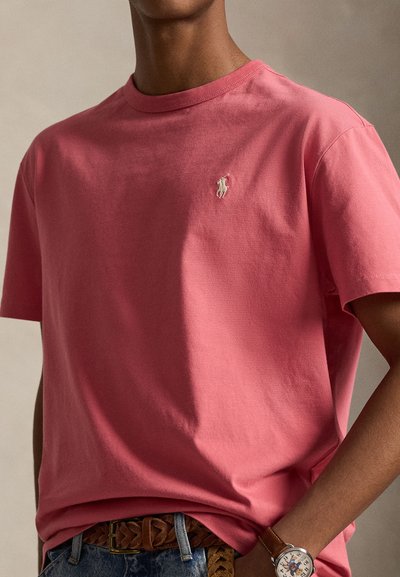 Άτομο που φοράει ροζ πόλο Ralph Lauren, καφέ πλεγτή ζώνη, τζιν και ρολόι χειρός με χαρακτήρα κόμικ στο καντράν.