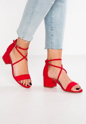 Czerwone sandały na obcasie z blokiem, z krzyżującymi się paskami w okolicy kostki, miękką tkaniną w górnej części oraz otwartym noskiem.