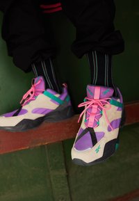 Personne portant des baskets violet, beige et turquoise avec des lacets roses et des chaussettes noires à rayures, assise les pieds pendant au-dessus d'un rebord en bois.