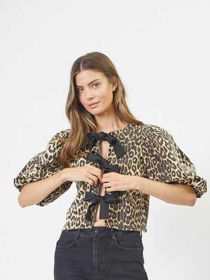 Blusa cropped con stampa leopardata, caratterizzata da maniche a sbuffo, un'apertura frontale a chiave con dettagli in legno neri, realizzata in tessuto morbido. Abbinata a jeans neri.