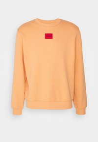 Bluza w kolorze brzoskwiniowym z okrągłym dekoltem, ściągaczami na mankietach i na dole. Na przodzie znajduje się czerwona prostokątna naszywka z logo "HUGO" w czarnym kolorze.