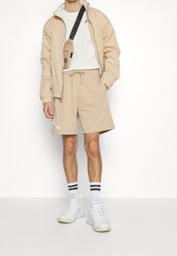 Beige lättviktsjacka med vita detaljer, åtföljd av beige shorts, vita sneakers och svartrandiga strumpor. Avslappnad sportklädsel.