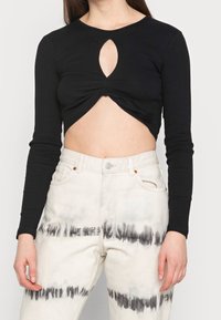 Top nero corto con maniche lunghe, caratterizzato da un'apertura circolare al centro e un dettaglio a torsione nella parte frontale. Abbinato a jeans crema con effetto tie-dye.