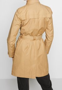 Beige trench coat kanssa vyötäröltä säädettävä, pitkät hihat ja takana tuuletusaukko. Sujuva, kevyt kangas rakenteellisella muotoilulla.