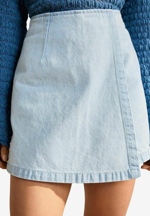 Mini-jupe en denim bleu clair portée avec un haut à manches longues bleu texturé, laissant apparaître le milieu de la cuisse et une partie des mains sur les côtés.