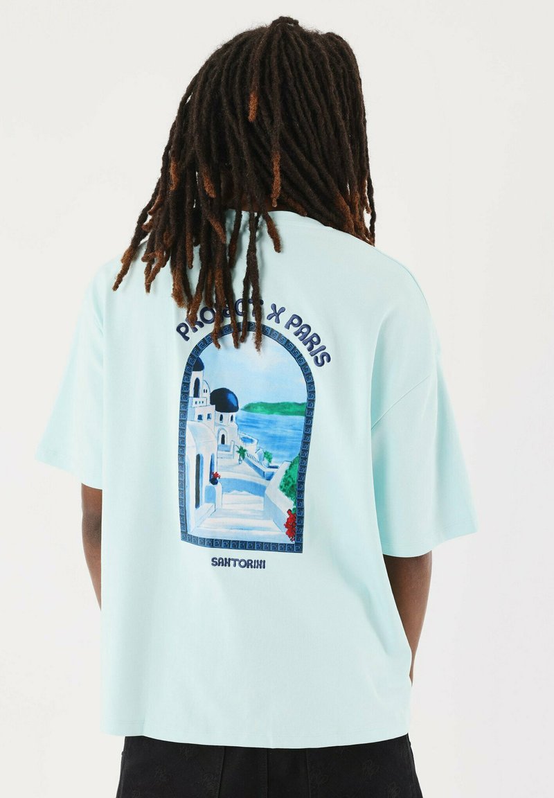 Camiseta de algodón de color azul claro con un diseño gráfico de Santorini en la parte posterior, con texto negro y elementos de paisaje detallados y coloridos.